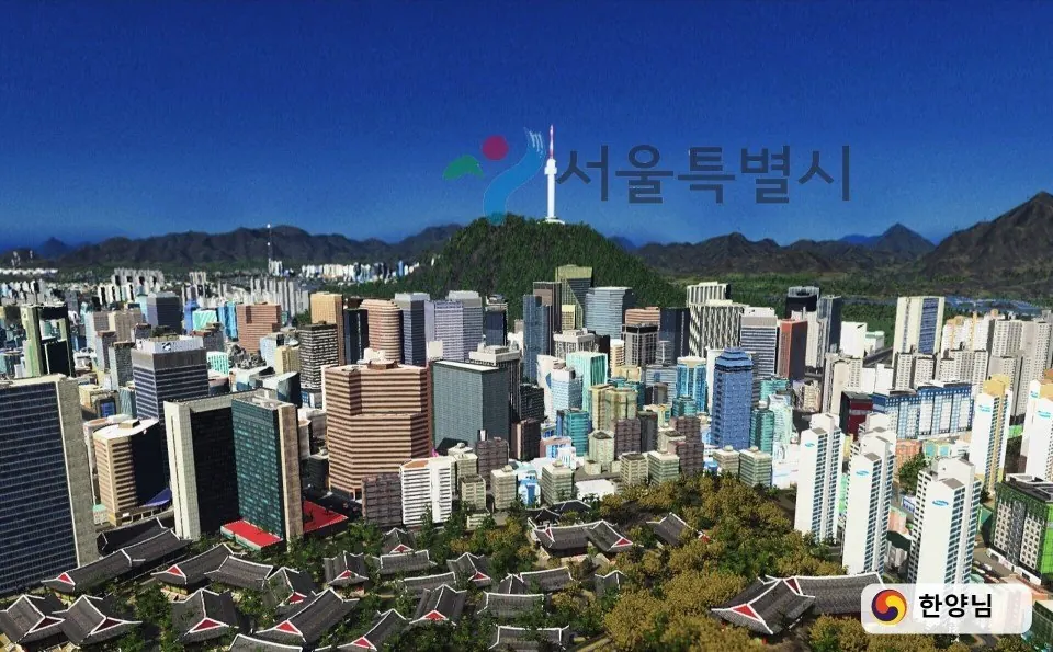 맑은 낮의 서울을 높은 곳에서 내려다본 화면. 앞쪽에 기와지붕의 한옥이 무리 지어 있고 그 뒤로 중고층 업무 건물이 빽빽이 이어지며, 가운데 녹색 산 위에 전망탑이 서 있다. 뒤로는 산줄기가 겹쳐 이어지고 하늘은 짙은 파랑이다.
