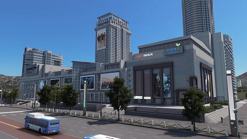대로변에 선 복합 문화·상업 건물. 벽면에 영화관과 서점 간판, 대형 포스터들이 걸려 있고 앞 인도에 미디어폴이 서 있다. 뒤로 고층 아파트와 산이 보이고 도로에는 파란 시내버스가 지난다.